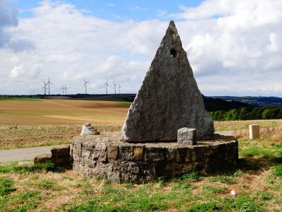 Das Bild zeigt eine pyramidenförmige Steinskulptur von Steine an der Grenze. Im Hintergrund sind Felder und Windräder zu sehen.