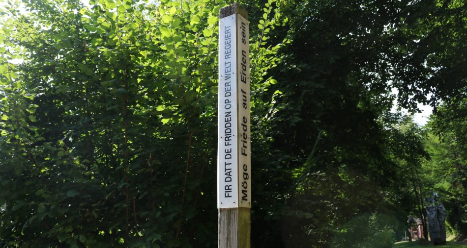 Das Foto zeigt einen Holzpfosten am Eingang des Parks der Andersdenkenden. Auf daran befestigten weißen Schildern ist rechts zu lesen: Möge Friede auf Erden sein. Links steht der gleiche Text auf luxemburgisch, der lautet: Fir datt de Fridden op der Welt regeiert.