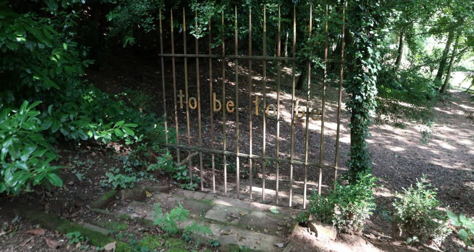 Das Bild zeigt die Installation von Herman de Vries im Park der Andersdenkenden. In einem Eisengitter mit goldenen Spitzen sind in Gold die englischen Begriffe to be to be eingesetzt.