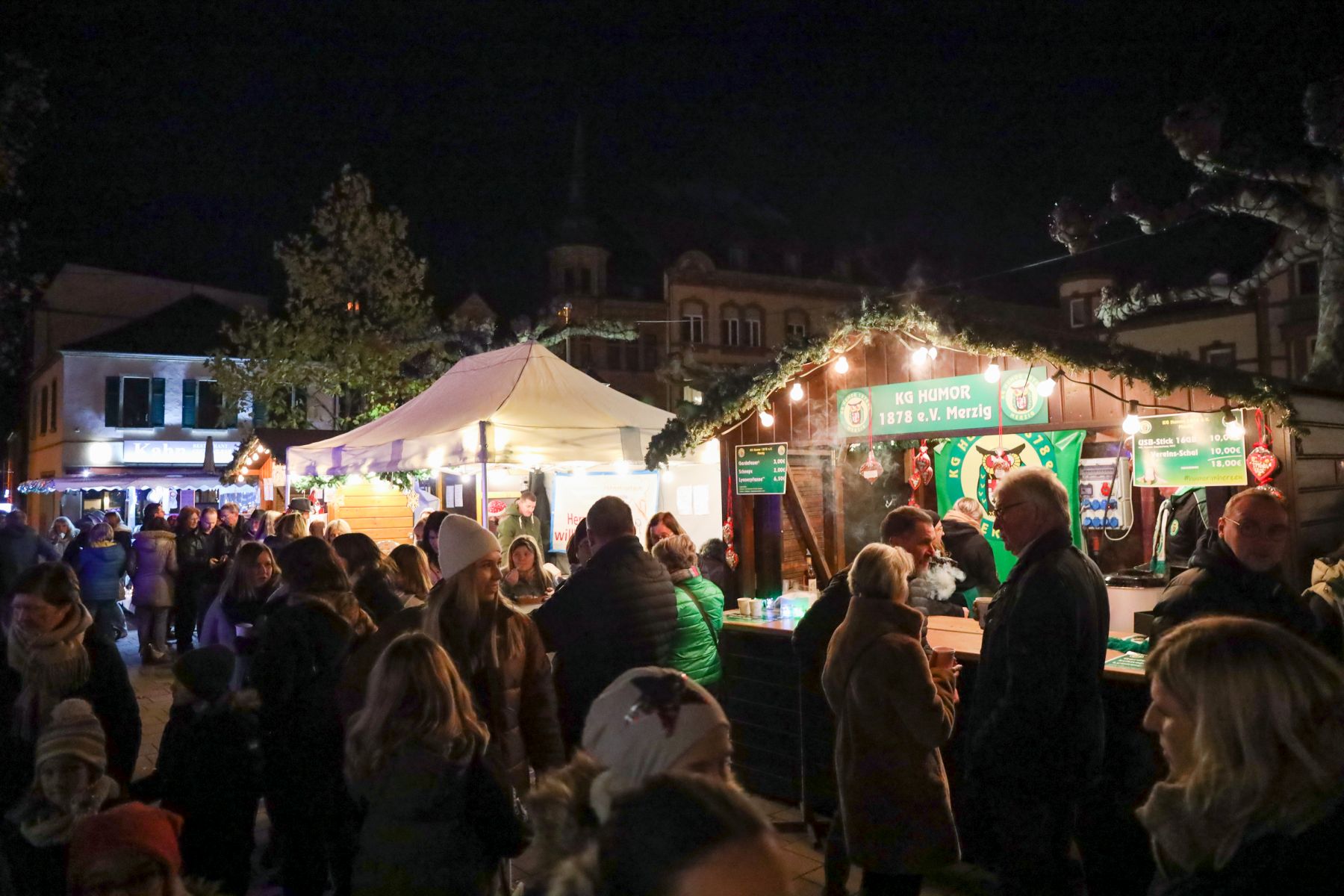Das Foto zeigt mehrere beleuchtete und weihnachtlich dekorierte Stände beim diesjährigen Nikolausmarkt. Zahlreiche Besucher sind ebenfalls zu sehen.