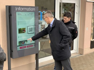 Das Foto zeigt das Info-Terminal am Wolfspark-Werner-Freund. Der Bürgermeister der Kreisstadt Merzig, Marcus Hoffeld steht davor und prüft gerade die Touch-Funktion des Terminals. Eine weitere Person steht neben ihm.