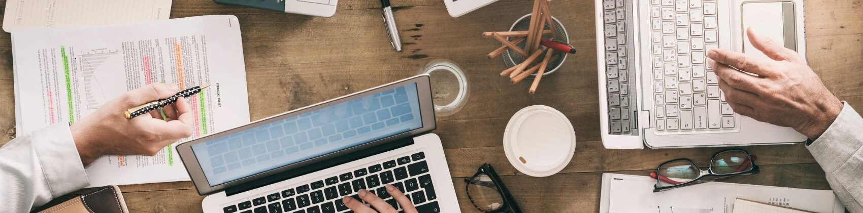 Stockfoto - Bild von oben zeigt Hände an Laptops, an einem Tablet, an einem Telefon, darum liegen Organizer, Stifte, Notizen
