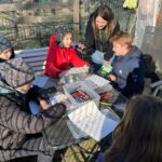 Fünf Kinder sitzen mit der Auszubildenden um einen Tisch, sie malen und beantworten Fragen auf Arbeitsblättern