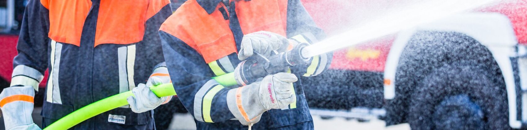 Oberkörper von zwei Feuerwehrkameraden in Uniform mit Schlauch in den Händen, aus dem Wasser spritzt