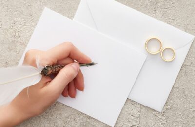 Zwei weiße Umschläge, zwei Ringe liegen auf einem Umschlag, eine Hand mit einem Füllfederhalter liegt auf dem zweiten Umschlag