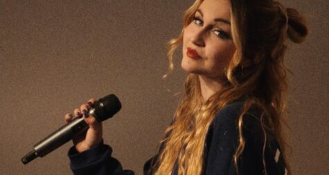 Elisa Rehlinger sitzt seitlich, ihr Kopf ist in Richtung Kamera geneigt, sie hält ein Mikro in der linken Hand