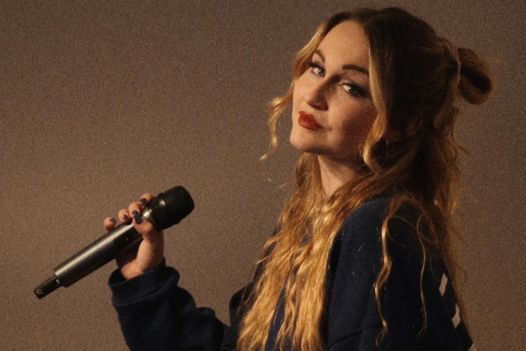 Elisa Rehlinger sitzt seitlich, ihr Kopf ist in Richtung Kamera geneigt, sie hält ein Mikro in der linken Hand