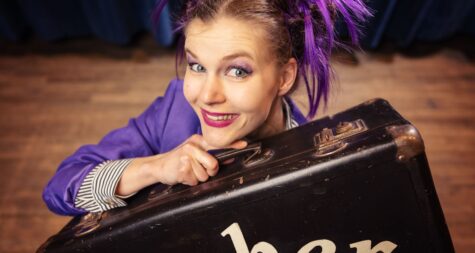 Frau mit lilafarbenen Strähnen lugt hinter einem Koffer hervor