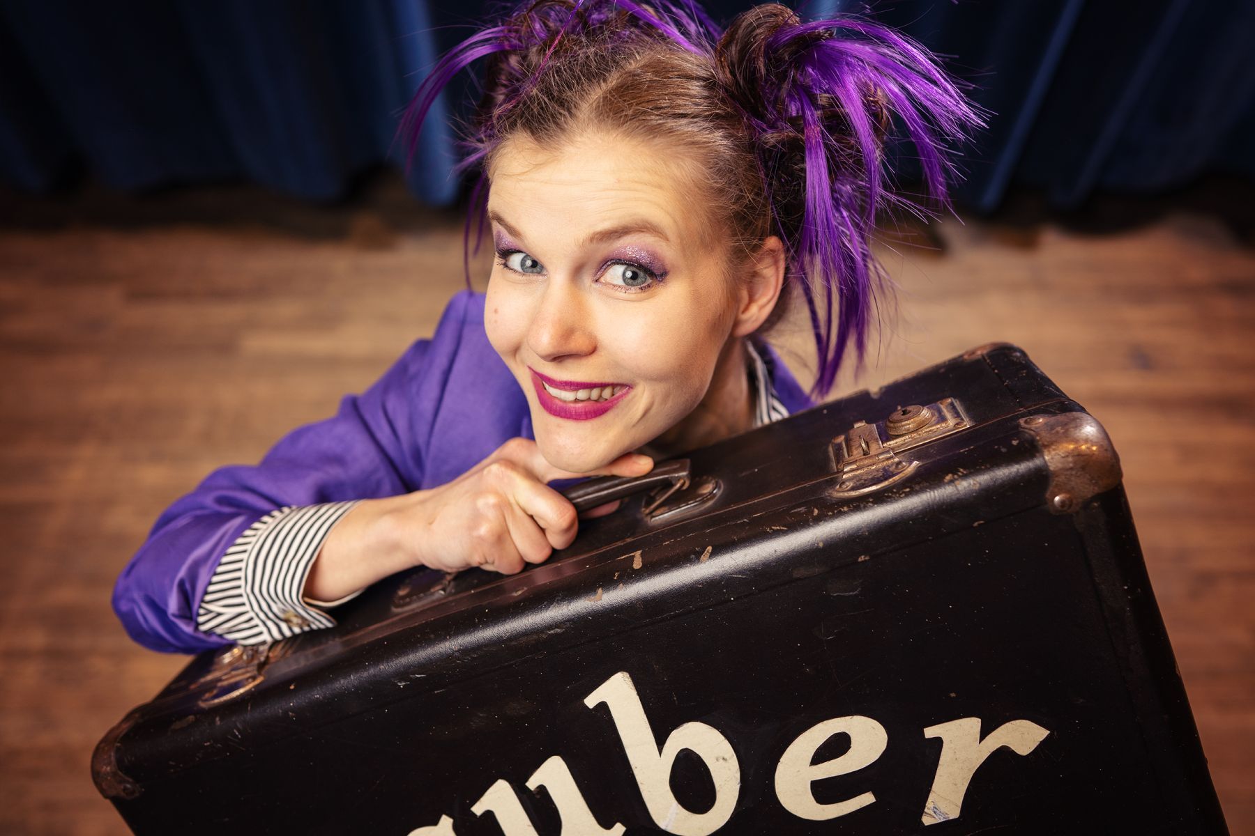 Frau mit lilafarbenen Strähnen lugt hinter einem Koffer hervor