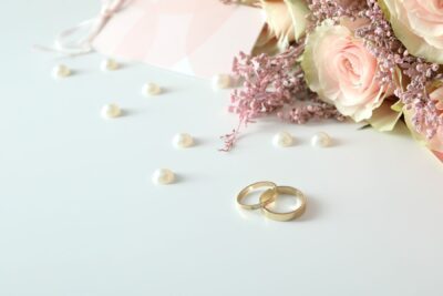 Zwei ineinander verschlungene Ringe liegen auf einem weißen Untergrund, daneben liegen helle Perlen, im rechten Rand sind rosa-weiße Rosen zu sehen