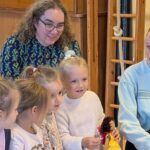 Vier Kinder sitzen, zwei Erwachsene knien auf dem Boden, ein Mädchen hält eine Handpuppe