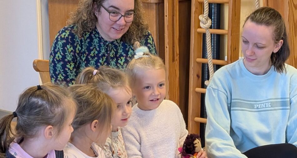 Vier Kinder sitzen, zwei Erwachsene knien auf dem Boden, ein Mädchen hält eine Handpuppe
