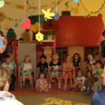 Kinder und Erwachsene in einem Kindergartenraum, Kinder sitzen auf kleinen Stühlen und hören einem Erwachsenen zu