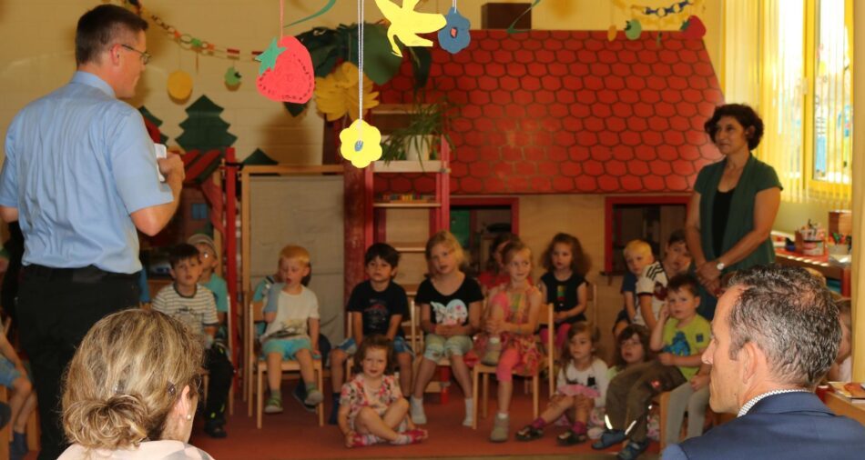 Kinder und Erwachsene in einem Kindergartenraum, Kinder sitzen auf kleinen Stühlen und hören einem Erwachsenen zu