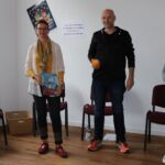 Vier Erwachsene, eine Frau hält ein Plakat, zwei Frauen halten Bücher, ein Mann hält eine Orange in der Hand