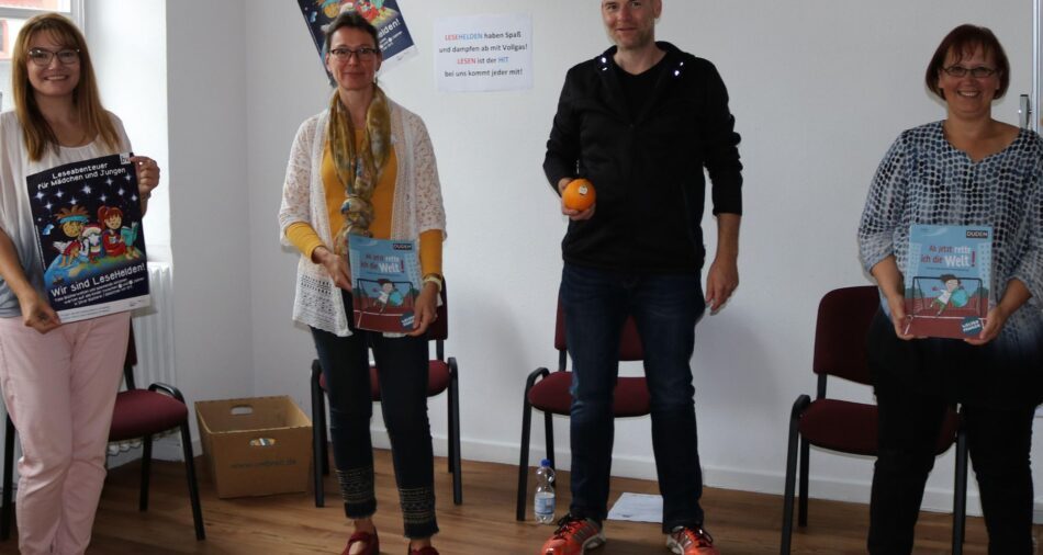 Vier Erwachsene, eine Frau hält ein Plakat, zwei Frauen halten Bücher, ein Mann hält eine Orange in der Hand