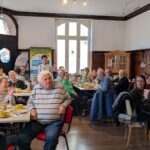 Menschen sitzen an Frühstückstischen, sie hören Oberbürgermeister Marcus Hoffeld zu