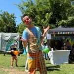 Mann mit buntem Clownkostüm posiert auf einer Wiese, im Hintergrund basteln und malen Kinder und Frauen