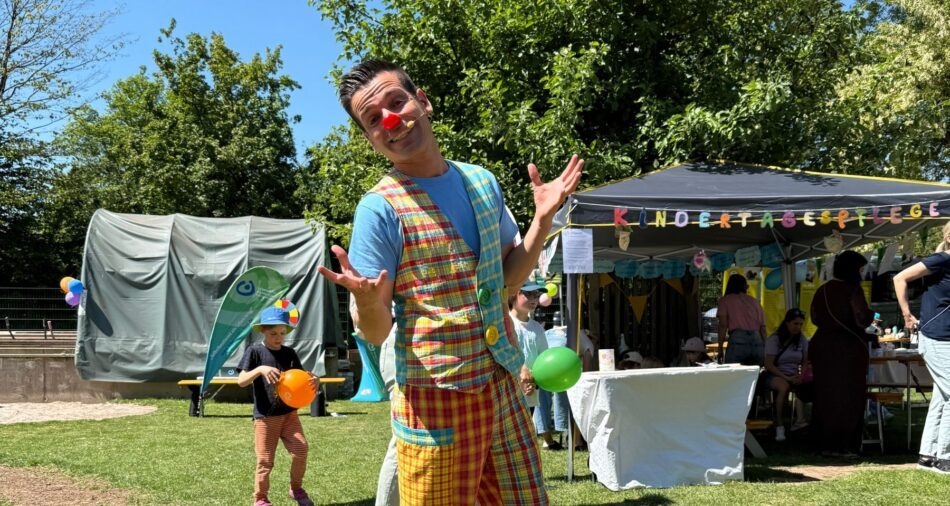 Mann mit buntem Clownkostüm posiert auf einer Wiese, im Hintergrund basteln und malen Kinder und Frauen