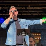 Mann singt auf einer Bühne, er hält in der rechten Hand ein Mikrofon, die linke Hand ist ausgebreitet