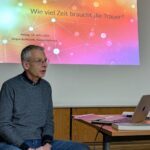 Mann sitzt vor einem Laptop, im Hintergrund eine Leinwand mit den Worten Wie viel Zeit braucht die Trauer