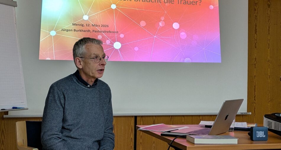 Mann sitzt vor einem Laptop, im Hintergrund eine Leinwand mit den Worten Wie viel Zeit braucht die Trauer