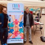 Zwei Männer posieren vor einem Banner, darauf steht Merzig miteinander