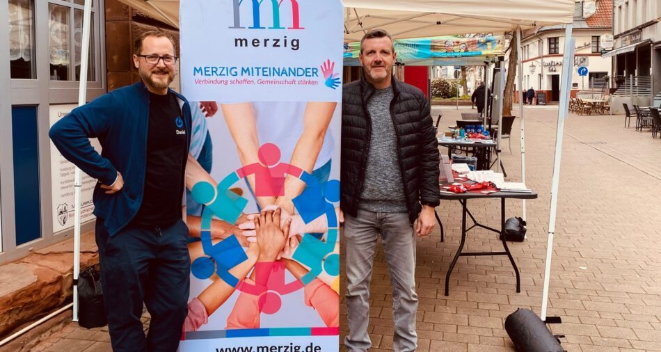 Zwei Männer posieren vor einem Banner, darauf steht Merzig miteinander
